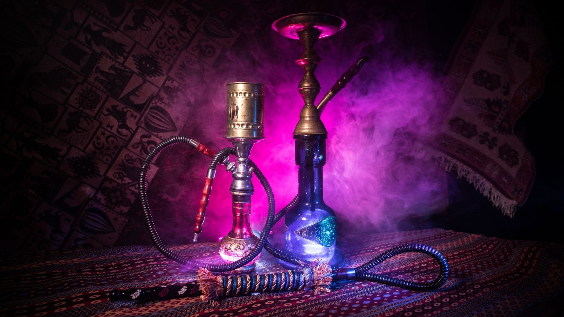 Two ornate hookahs with glowing blue and pink smoke on patterned fabric